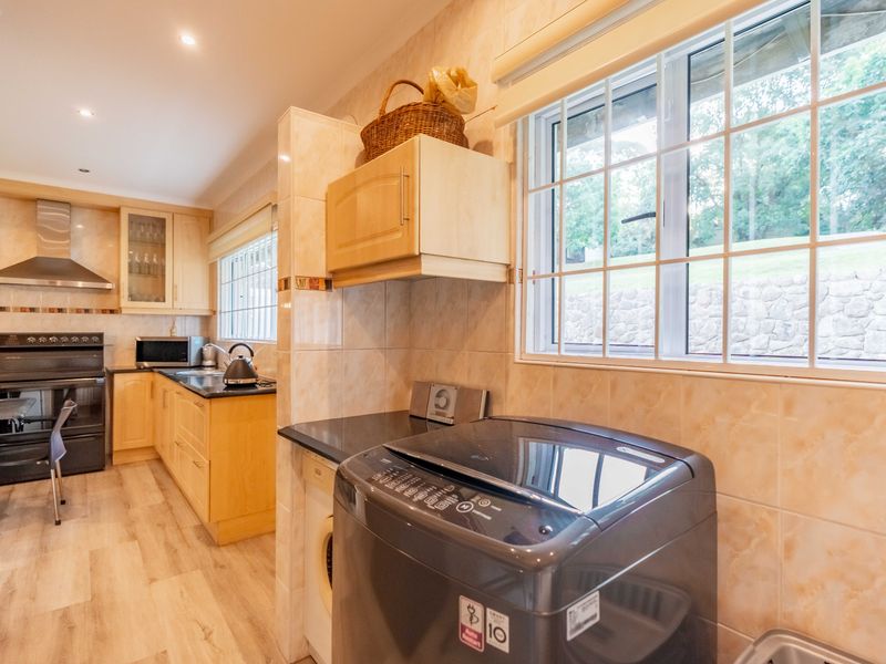 A functional kitchen featuring light wood cabinetry, dark countertops, integrated appliances, and a tiled splashback, with an adjacent laundry area.