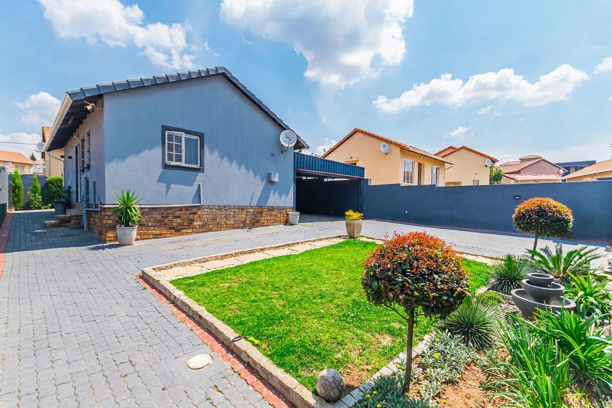 A well-maintained rear exterior view featuring a paved driveway, a covered carport, and a neat landscaped garden with a brick boundary wall.