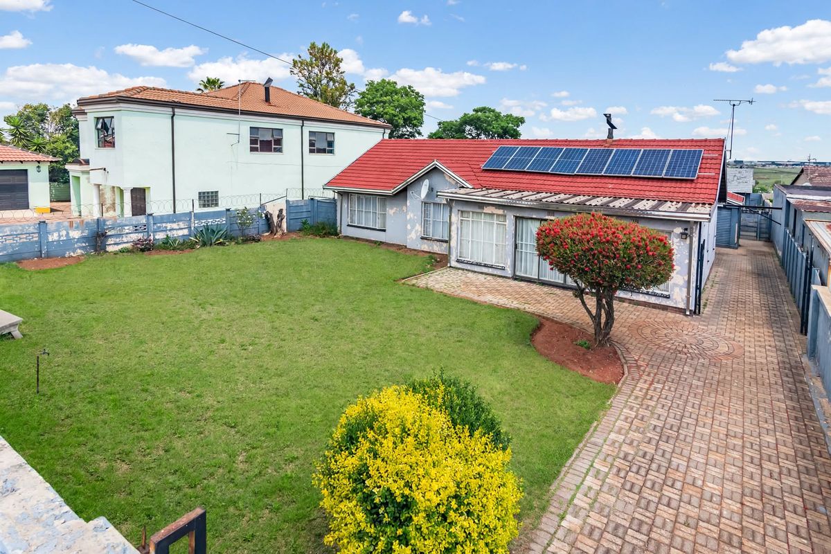 Rear view of the property featuring a spacious lawn, paved patio area, and roof-mounted solar panels, with visible wear to the exterior wall paint.