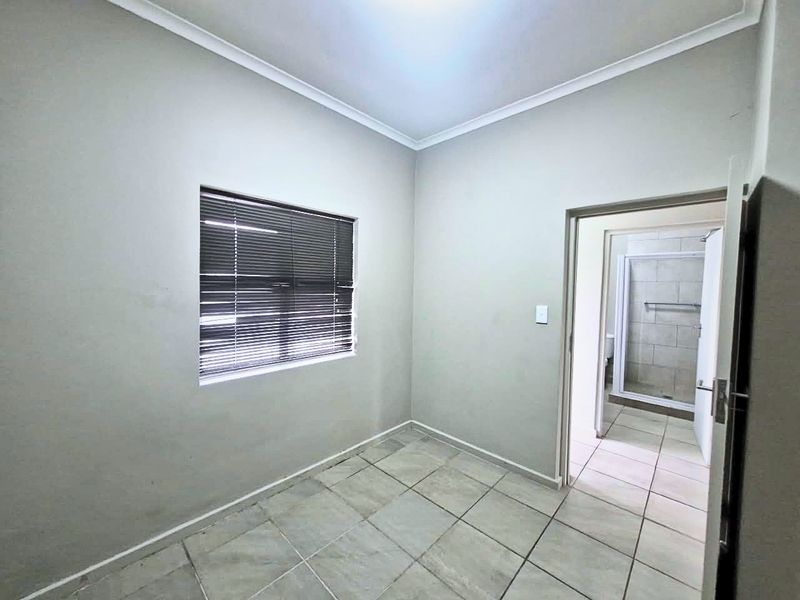 An empty bedroom featuring neutral tiled flooring, coving, and a window fitted with venetian blinds, with an adjoining bathroom visible through the doorway.
