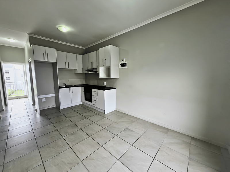 A clean, empty kitchen featuring white cabinetry, dark countertops, built-in oven, extractor fan, and tiled flooring, leading to an adjacent balcony.