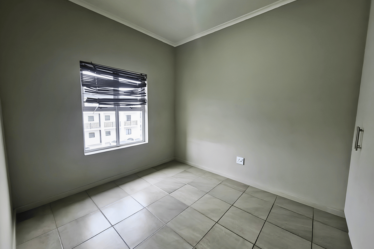 An empty bedroom featuring neutral tiled flooring, a built-in cupboard, and a window fitted with venetian blinds.