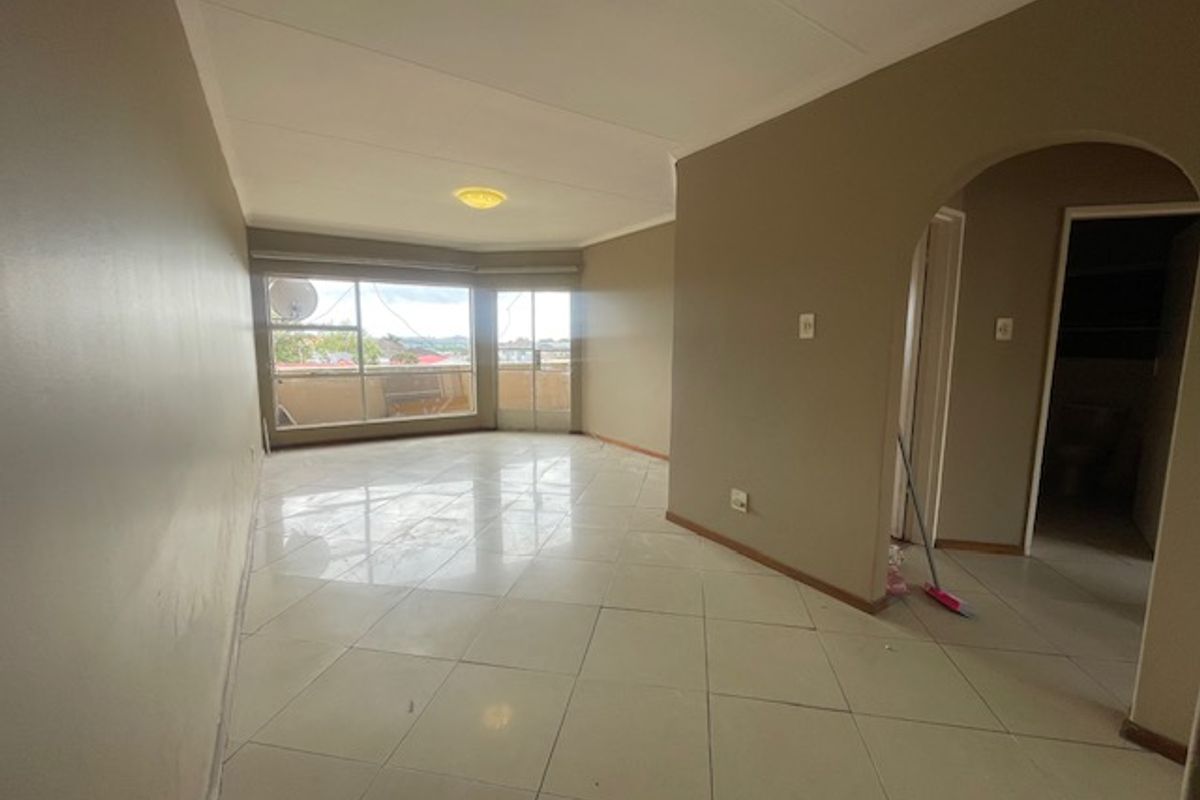 A spacious, empty living area featuring neutral tiled flooring, an arched entryway, and large windows providing natural light and access to a balcony.