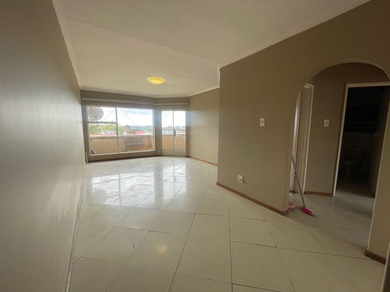 A spacious, empty living area featuring neutral tiled flooring, an arched entryway, and large windows providing natural light and access to a balcony.