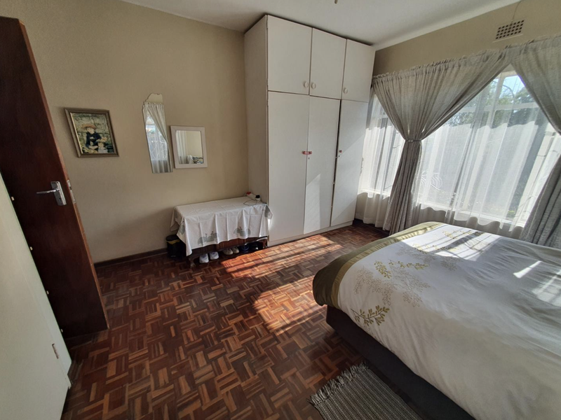 A bedroom featuring classic parquet flooring, built-in cupboards, and large windows with curtain rails, offering a functional and well-lit living space.