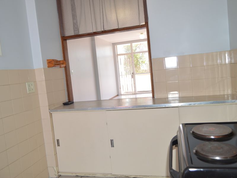 A dated kitchen featuring built-in cabinetry, a metal countertop, and a tiled splashback, with a serving hatch opening into the adjacent living area.