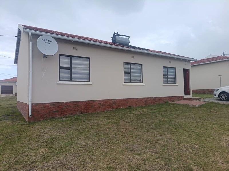 A single-story residential house featuring a tiled roof, solar water heating system, and brick plinth detailing, set on a grassed plot.