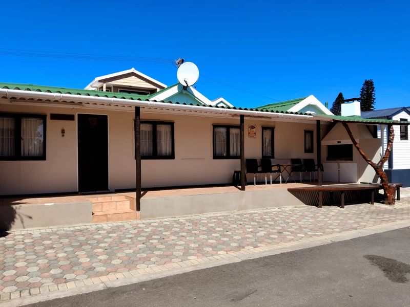 Front exterior view of a single-story house featuring a covered patio, built-in braai area, and paved driveway under a clear blue sky.