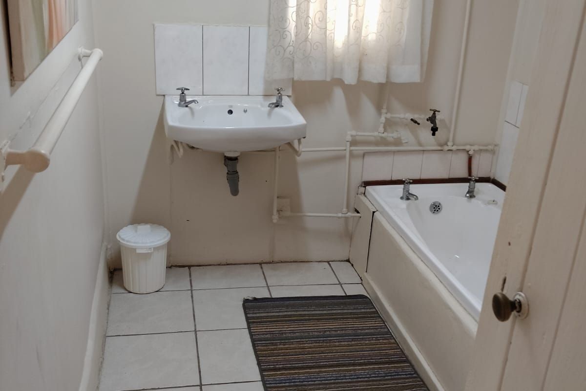 A compact bathroom featuring a wall-mounted basin, bathtub, and tiled flooring, with visible surface-mounted plumbing and dated finishes throughout.