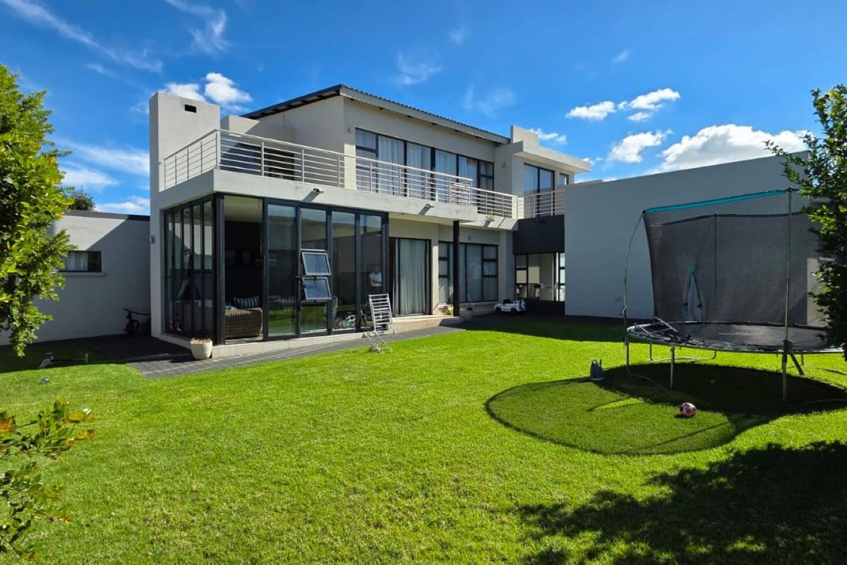 A spacious rear exterior view of a modern two-story house featuring a large lawn, paved patio, and an upper-level balcony with metal railings.