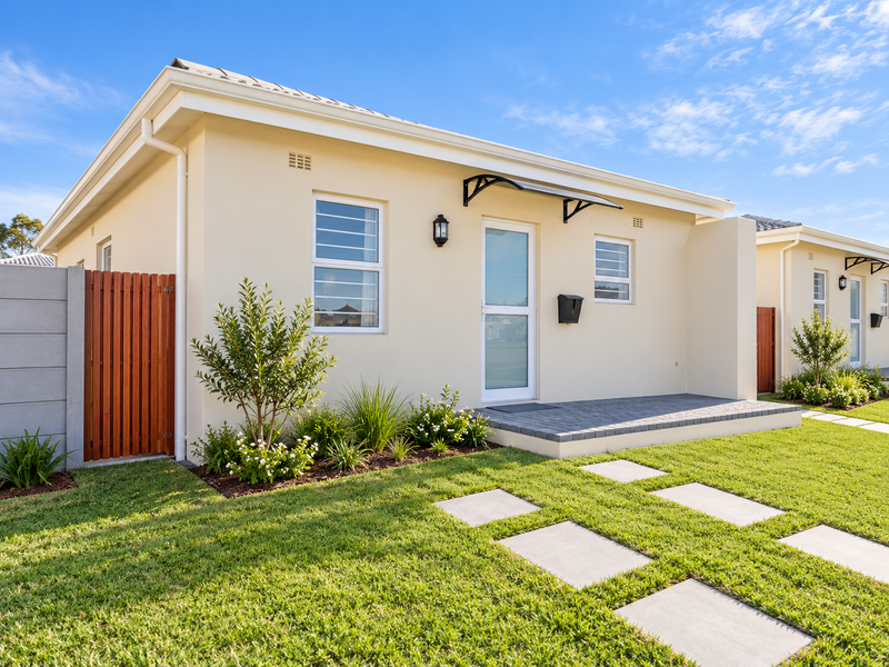A well-maintained modern single-story home exterior featuring a paved walkway, landscaped garden, timber gate, and a covered entrance area.