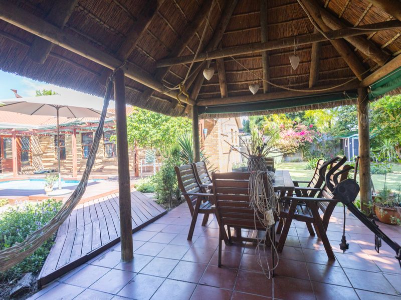 A covered outdoor patio area featuring a traditional thatched roof, timber support posts, and tiled flooring, overlooking a garden and swimming pool.