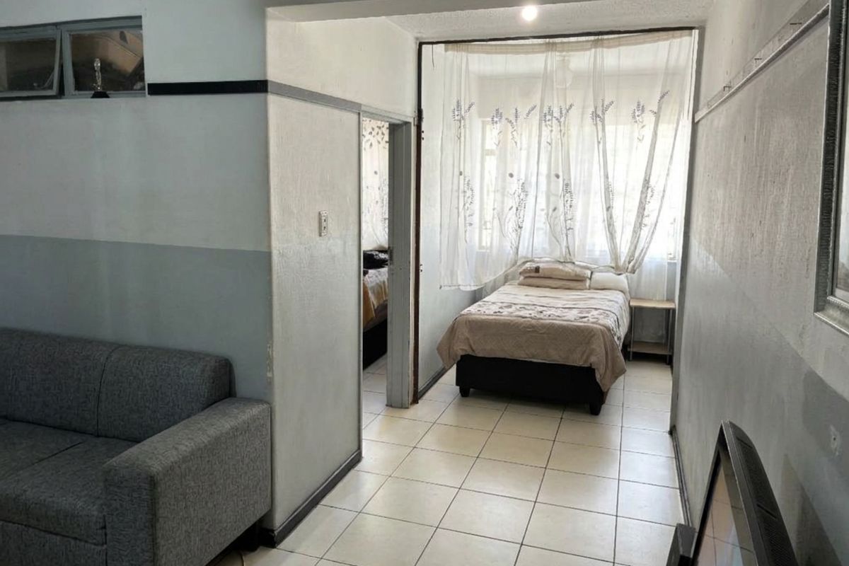 A living area featuring tiled flooring and a view into an adjacent bedroom, showing dated wall finishes and basic structural layout.