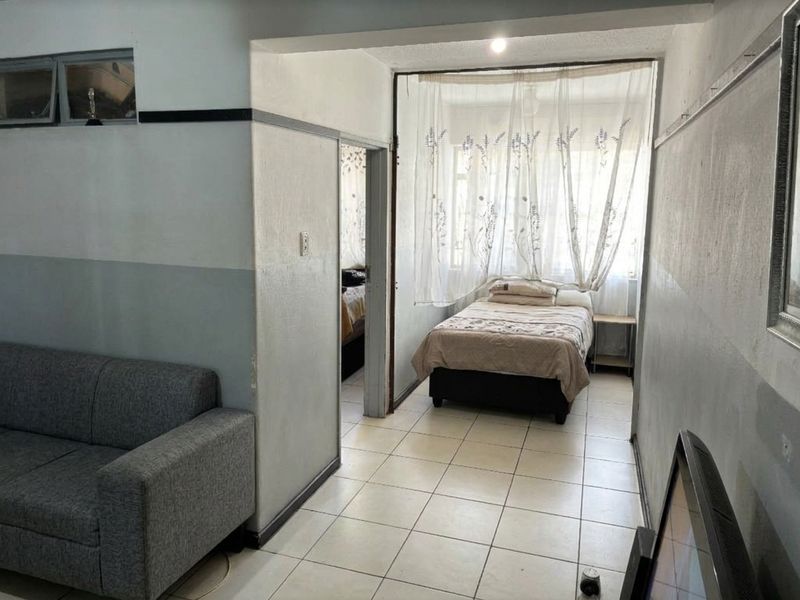 A living area featuring tiled flooring and a view into an adjacent bedroom, showing dated wall finishes and basic structural layout.
