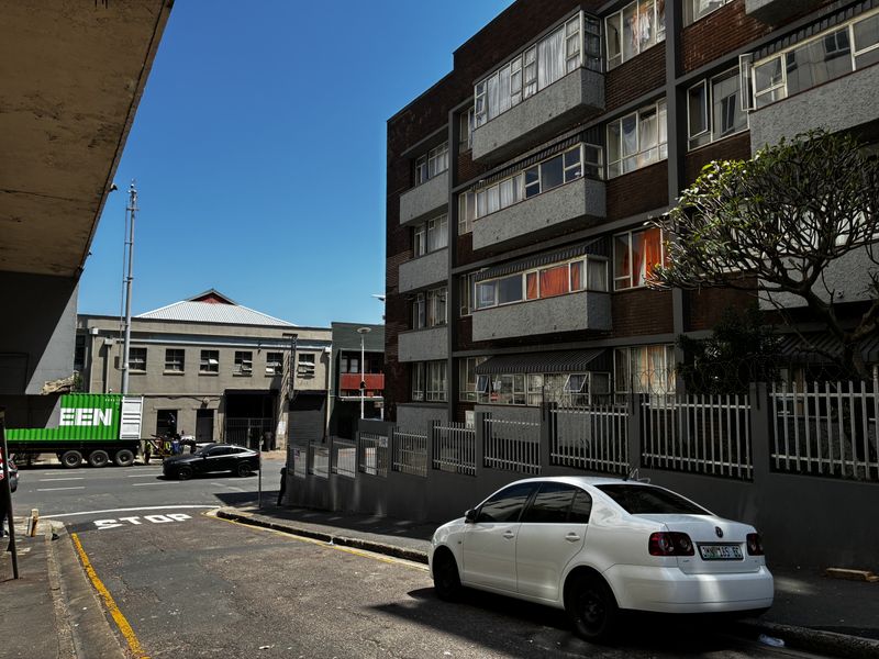 Street view of a multi-story brick apartment building with balconies and perimeter security fencing in an urban setting.