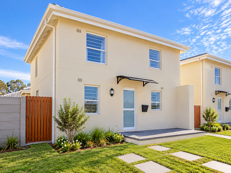 A modern two-story residential facade featuring cream-colored exterior walls, a paved entrance area, and a well-maintained front garden with a timber side gate.