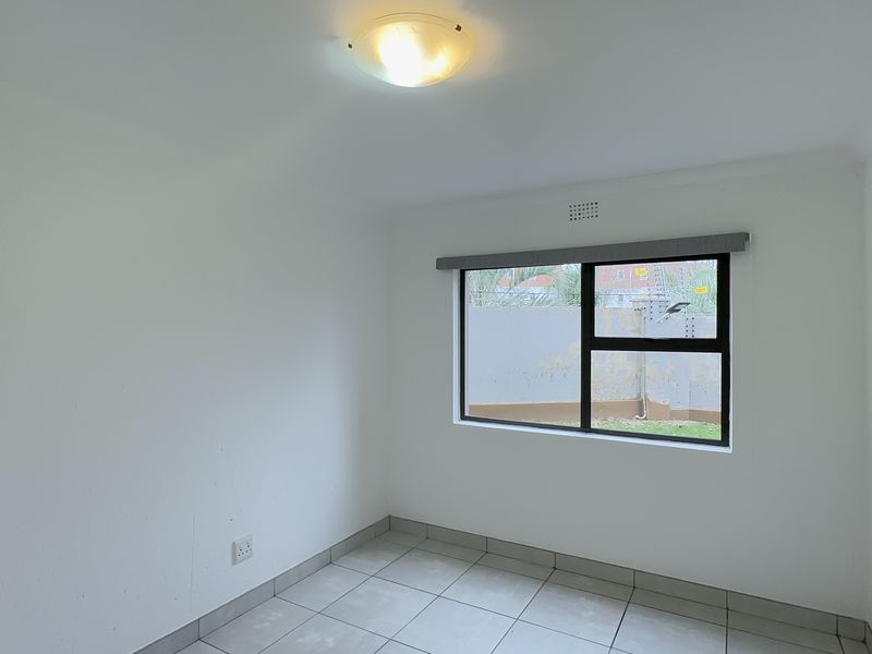 A clean, empty bedroom featuring neutral tiled flooring, white walls, and an aluminium-framed window providing natural light.
