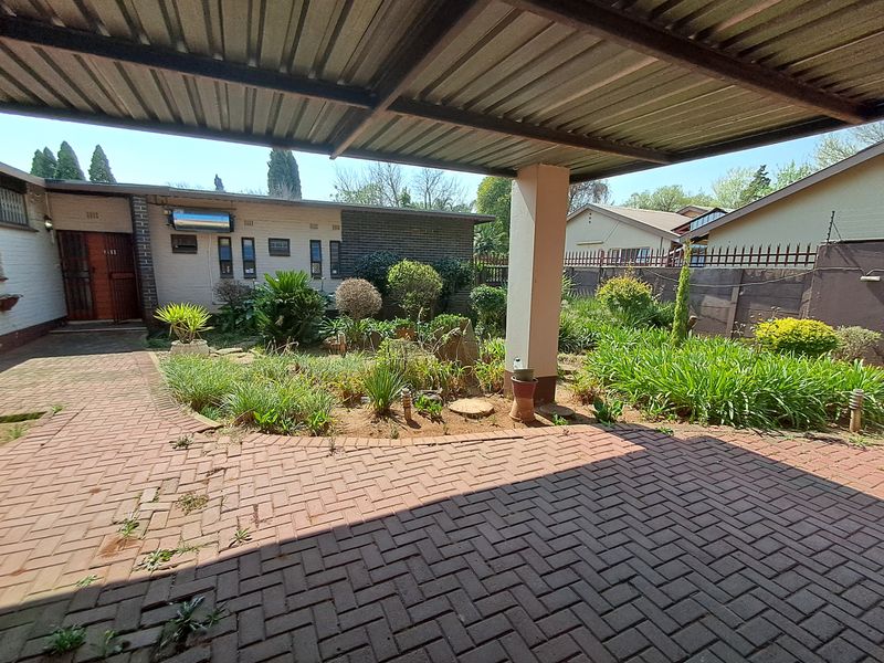 A covered brick-paved patio area overlooking a landscaped garden, featuring a corrugated metal roof structure and direct access to the main building.