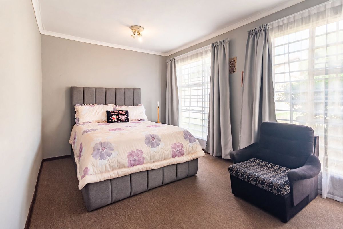 A well-lit bedroom featuring neutral wall tones, carpeted flooring, and large windows fitted with security bars.