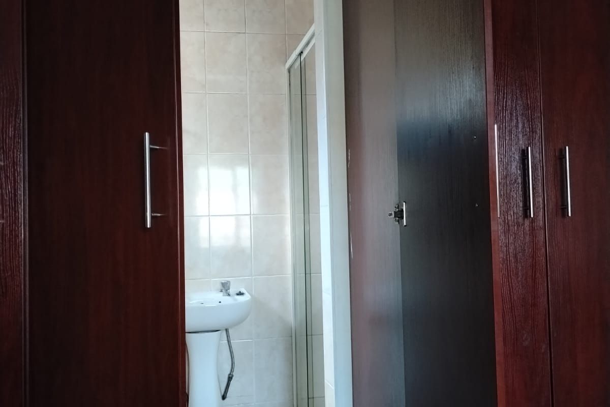 A functional bathroom featuring a pedestal basin, shower enclosure, and neutral wall tiling, accessed via a hallway with built-in wooden cabinetry.