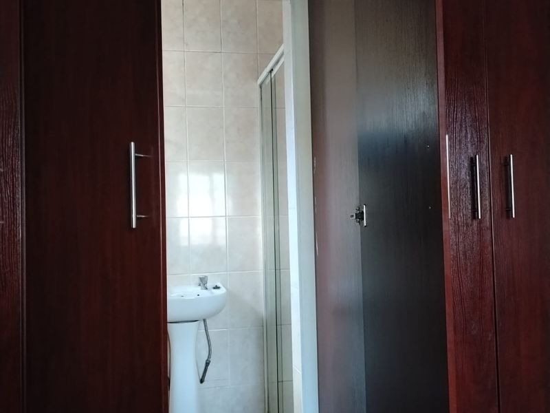 A functional bathroom featuring a pedestal basin, shower enclosure, and neutral wall tiling, accessed via a hallway with built-in wooden cabinetry.