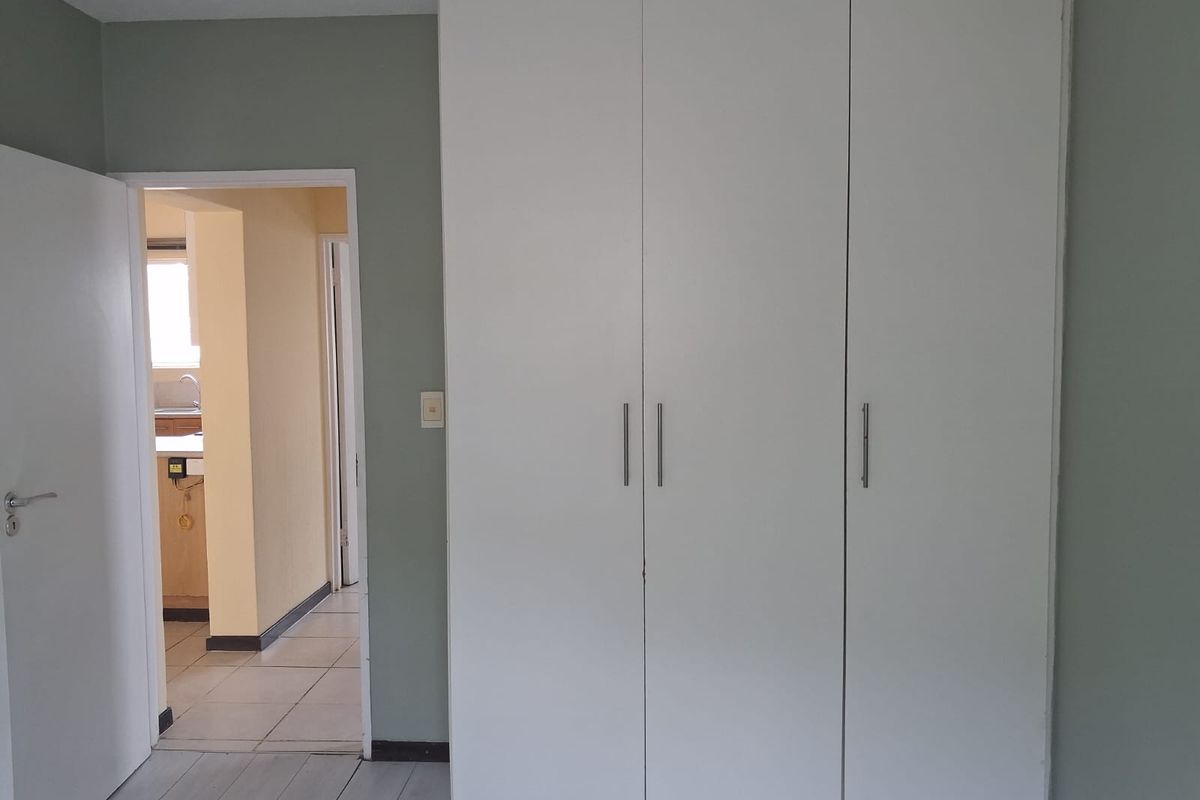 A bedroom featuring a large white built-in wardrobe, neutral wall paint, and tiled flooring, with a view into the adjacent kitchen area.