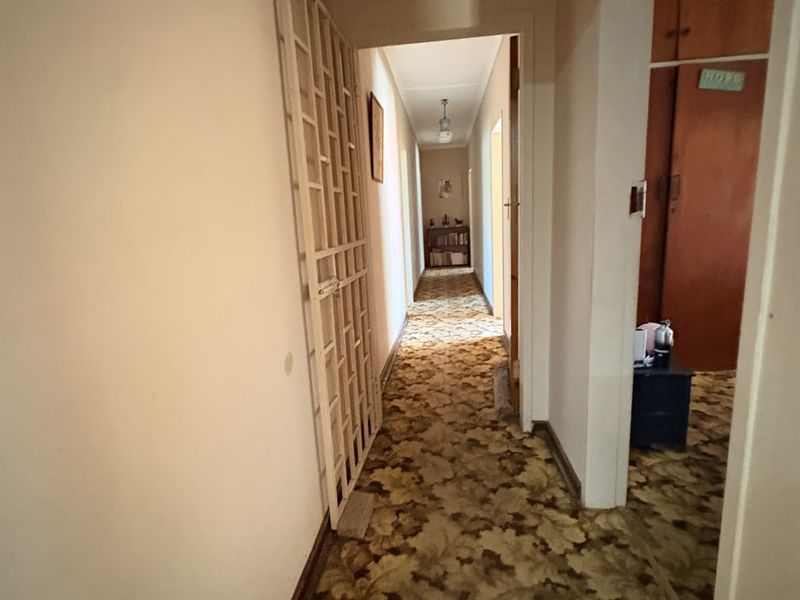 A long residential hallway featuring dated floral carpet flooring, a security gate, and built-in wooden cupboards.