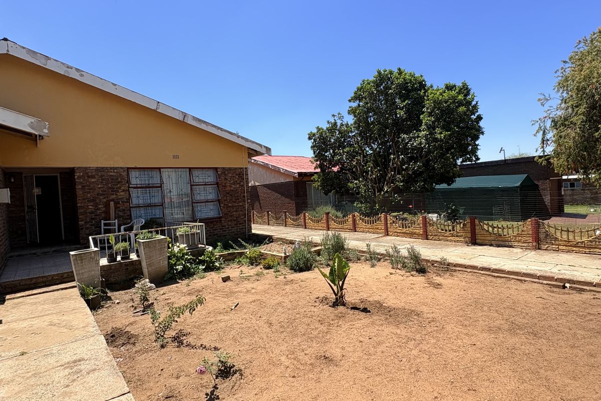 Rear view of the property featuring a brick and plaster facade, concrete walkway, and a large open garden area with perimeter fencing.