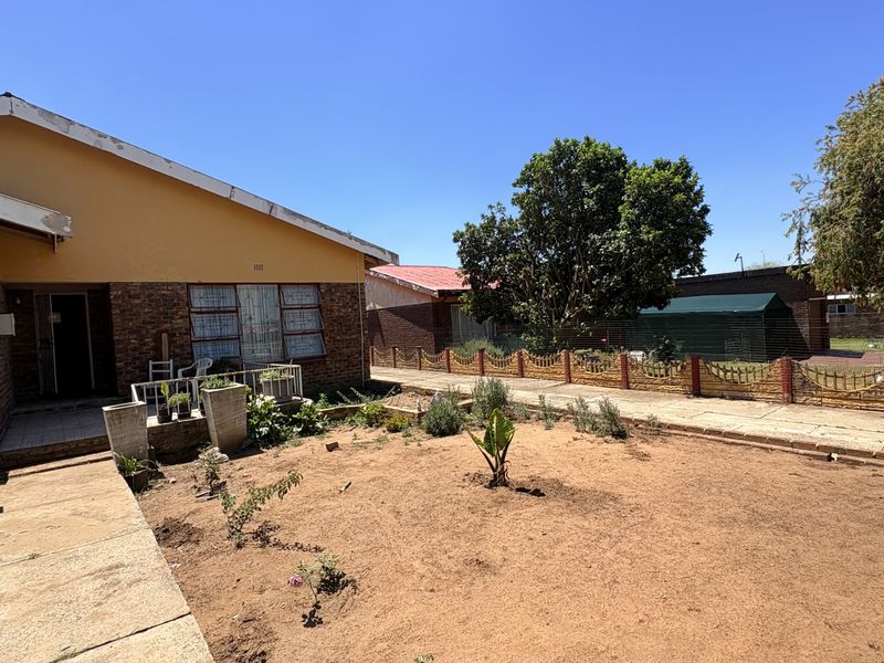 Rear view of the property featuring a brick and plaster facade, concrete walkway, and a large open garden area with perimeter fencing.