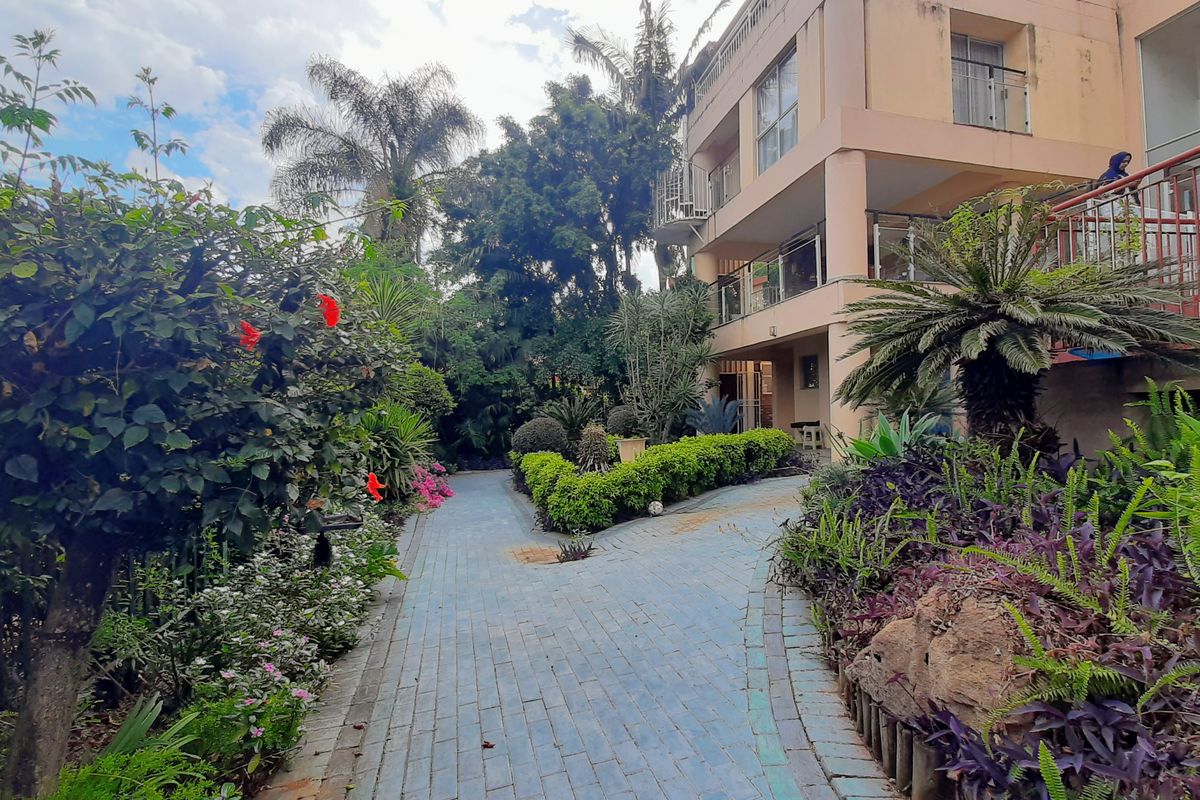 A view of the rear garden area featuring a paved walkway leading towards a multi-story residential building with balconies and established landscaping.