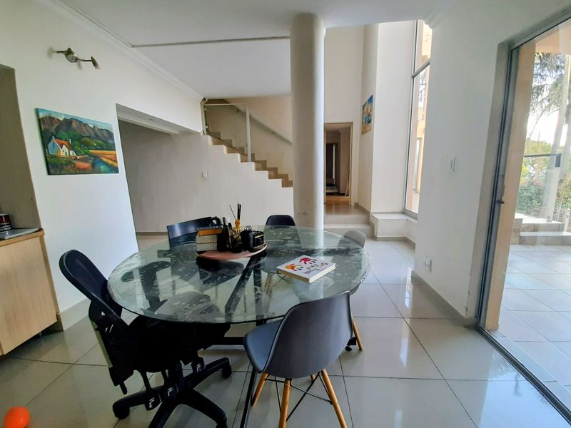 Bright dining area featuring large format floor tiles, a central structural column, and a modern staircase with glass balustrade leading to upper levels.