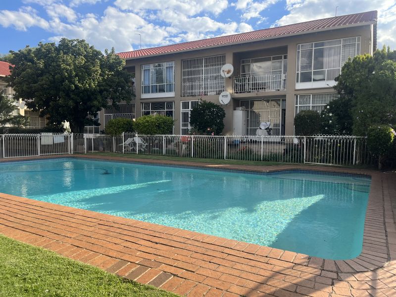 A well-maintained communal swimming pool area featuring brick coping and metal fencing, set against the rear facade of a multi-story apartment building.