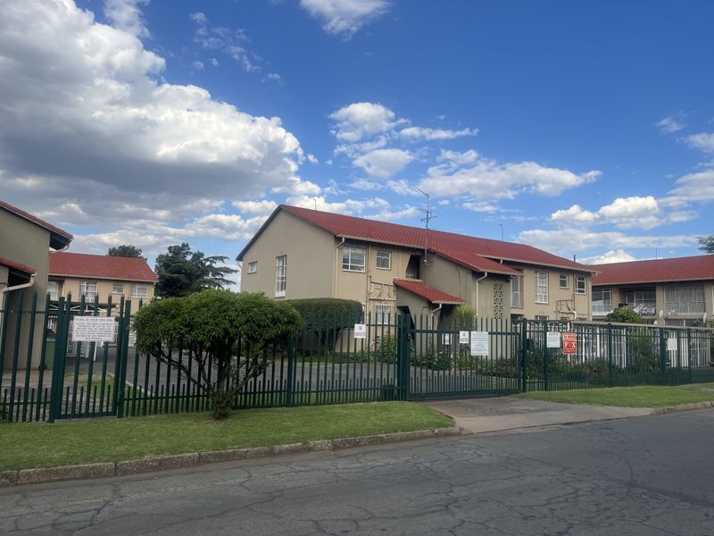 Street view of an apartment complex featuring a red tiled roof, paved driveway, and secure metal perimeter fencing.