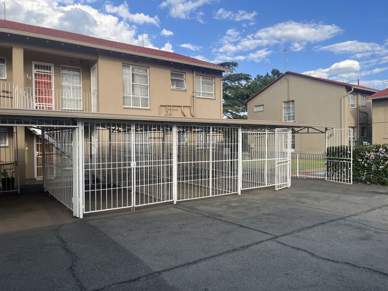 Exterior view of an apartment building featuring a covered carport with security gates and a paved driveway area.