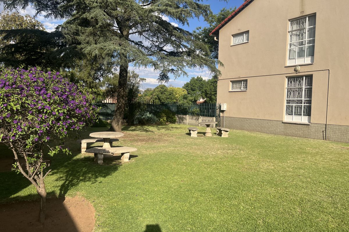 A well-maintained communal garden area featuring a lawn, mature trees, and security-fitted windows on the rear facade of the apartment building.