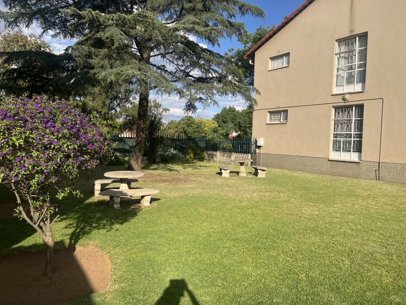 A well-maintained communal garden area featuring a lawn, mature trees, and security-fitted windows on the rear facade of the apartment building.