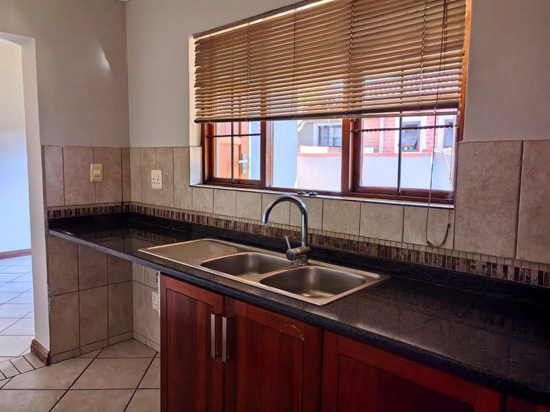 Kitchen area featuring granite countertops, double bowl stainless steel sink, wooden cabinetry, and tiled backsplash with natural light from a window with venetian blinds.