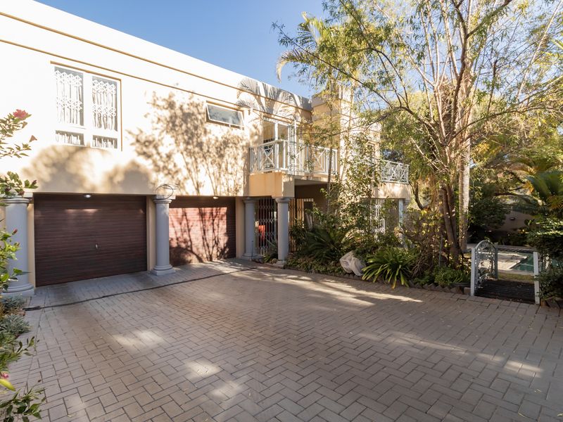 Front elevation of a duet property featuring a paved driveway, double garage, and an upper-level balcony with established garden surroundings.