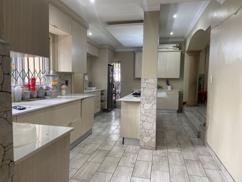 Modern kitchen featuring light wood cabinetry, stone countertops, a central island, and tiled flooring, with an open-plan layout leading to an adjacent living area.
