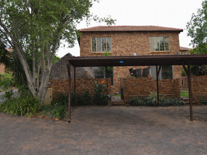 Front exterior view of a double-story brick townhouse featuring a paved driveway, a covered carport, and established garden landscaping.