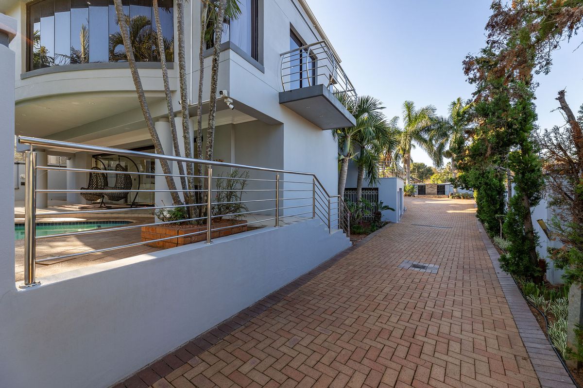 A well-maintained rear exterior view featuring a paved driveway, stainless steel balustrades, a balcony, and a swimming pool area.