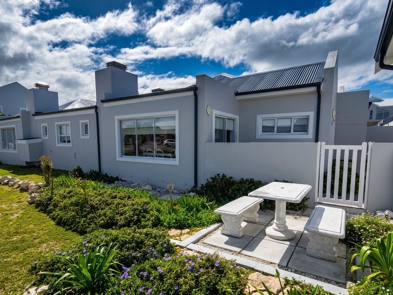 Rear view of a modern apartment featuring a paved patio, landscaped garden, white picket gate, and clean grey exterior walls with sliding windows.