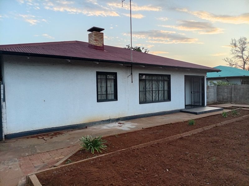 Rear view of a single-story house featuring a corrugated metal roof, brick chimney, security-fitted windows, and a concrete patio overlooking a garden area.