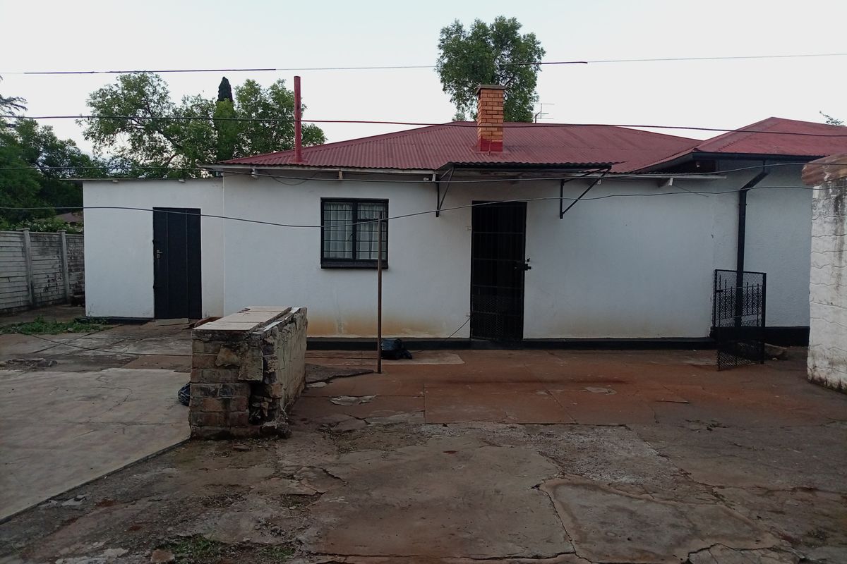 Rear exterior view of a house featuring a corrugated metal roof, brick chimney, and a concrete patio area requiring maintenance and repair.