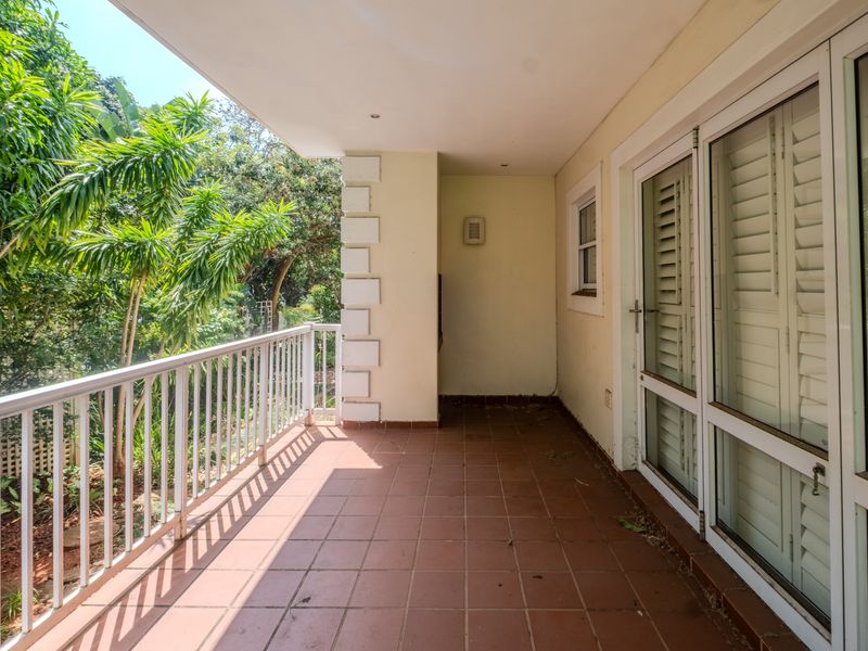 A covered balcony featuring tiled flooring, a metal balustrade, and sliding glass doors, overlooking a lush garden area.