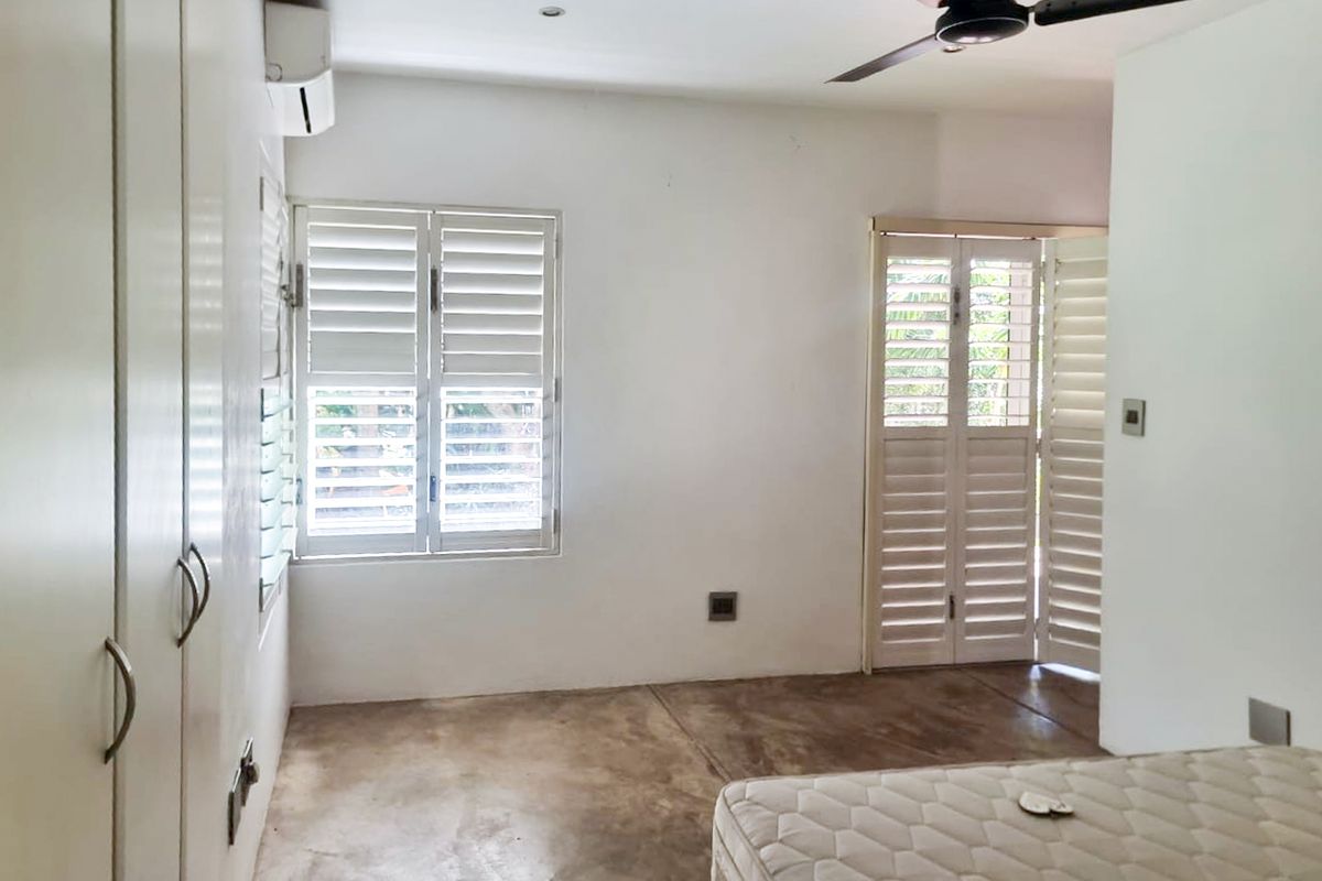 A bright, minimalist bedroom featuring polished concrete floors, built-in storage, plantation shutters, and an integrated air conditioning unit.