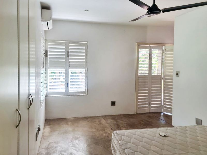A bright, minimalist bedroom featuring polished concrete floors, built-in storage, plantation shutters, and an integrated air conditioning unit.