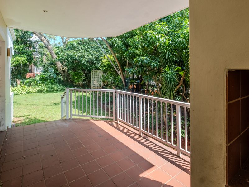 A covered patio featuring tiled flooring, a built-in braai, and a metal balustrade, overlooking a lush private garden.