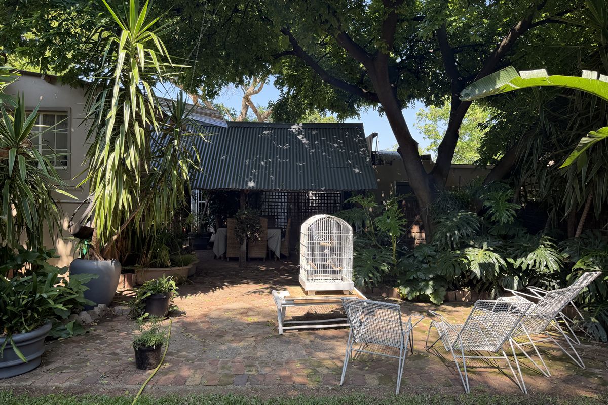 A shaded rear garden area featuring a brick-paved patio and a covered outdoor structure nestled under mature trees.