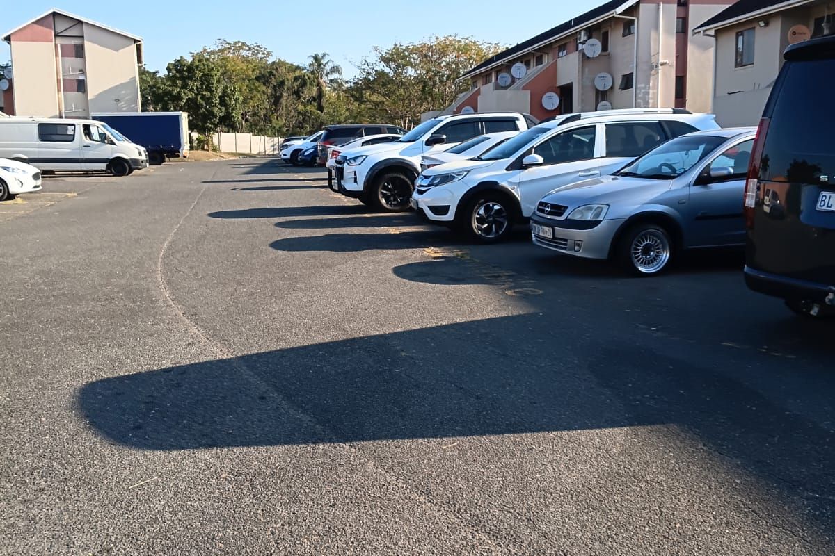 An outdoor parking area situated between multi-story apartment buildings, featuring paved surfaces and designated vehicle parking spaces.